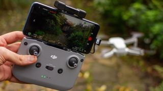DJI Mini 2-controller holdt i venstre hånd med drone i baggrunden