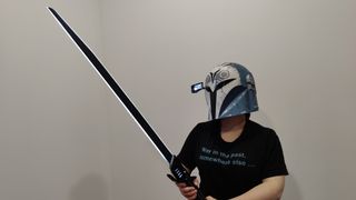 Person wearing the Black Series Bo-Katan helmet with the heads-up display down and lit up. They are holding the Darksaber (blade sword with white glowing edge) in an attack position and wearing a black t-shirt with light blue text that reads “Way in the past, somewhere else…”.