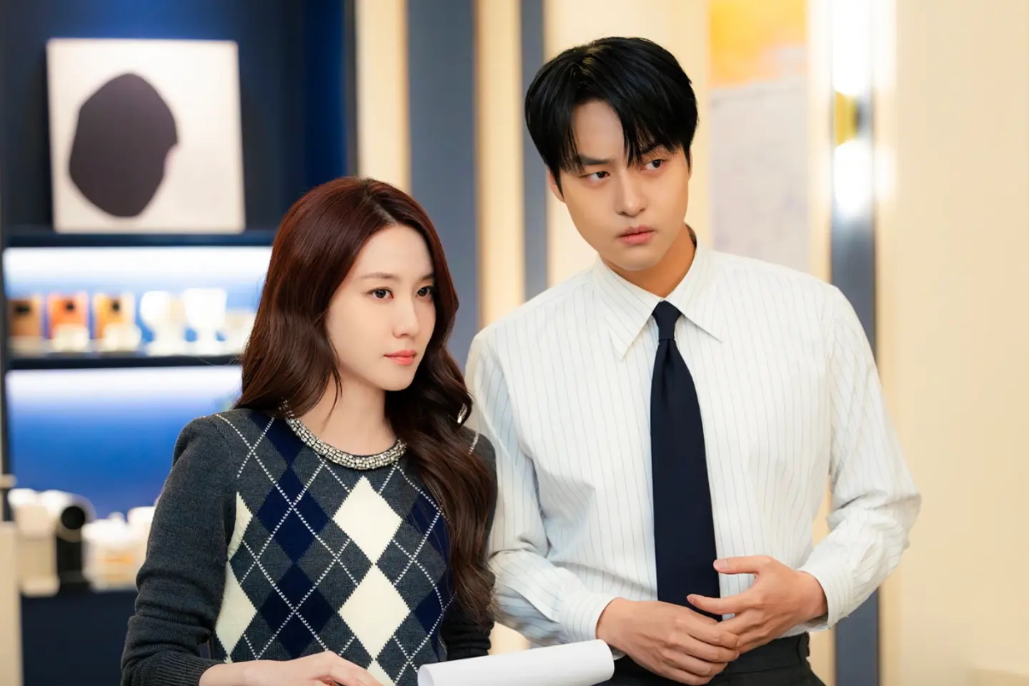 A woman in an argyle sweater (actress Park Eun-bin) and a man in a white shirt and tie (actorYang Se-jeong) stand in an office building, in the Korean drama 'Spooky in Love.'