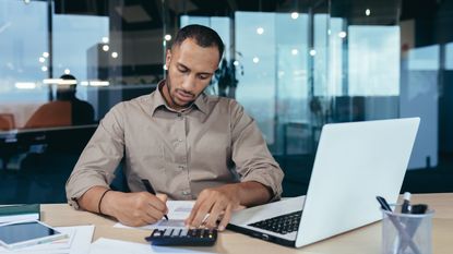 A man sits at a desk in an office and uses a calculator.