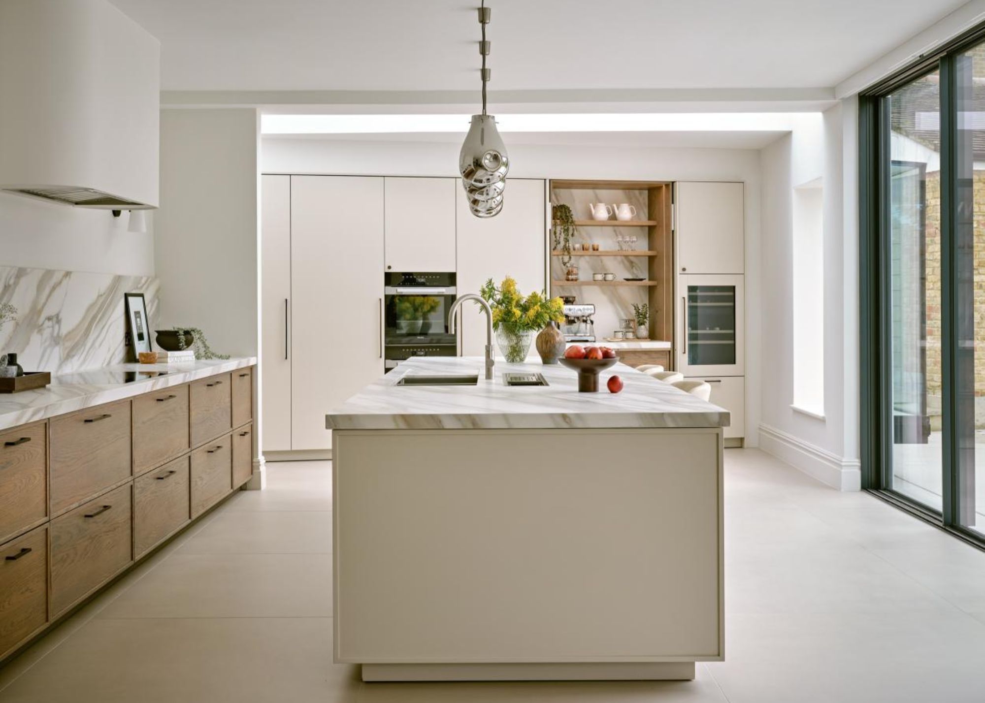 A neutral kitchen with chrome pendant lights above a large kitchen island in front of sliding glass doors with built-in cabinetry on the back wall with an alcove with decorative items