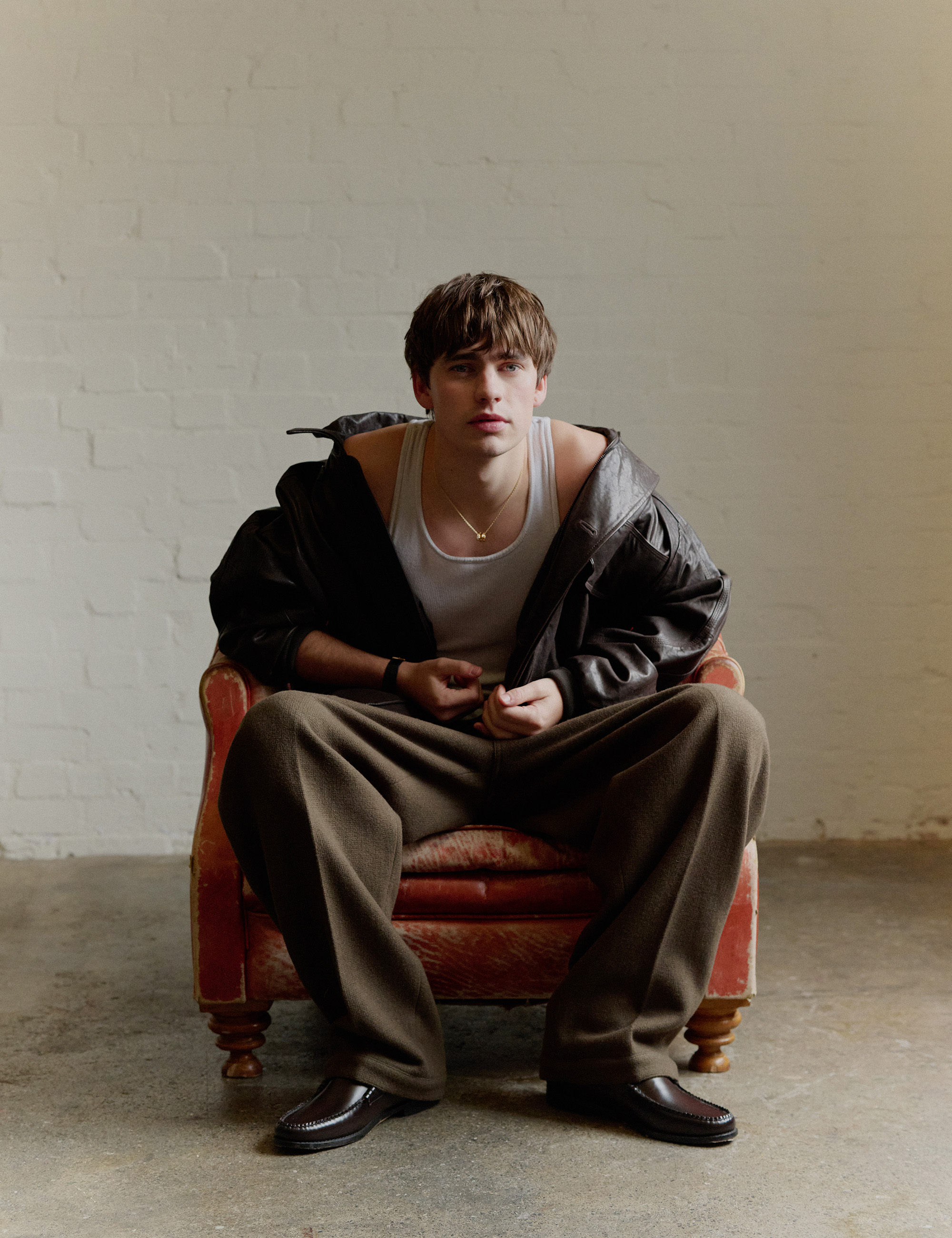 Spike Fearn sits in a worn orange leather chair against a white brick wall background. He is wearing a dark brown leather jacket over a white tank top, Bulgari necklace, brown trousers, and loafers.