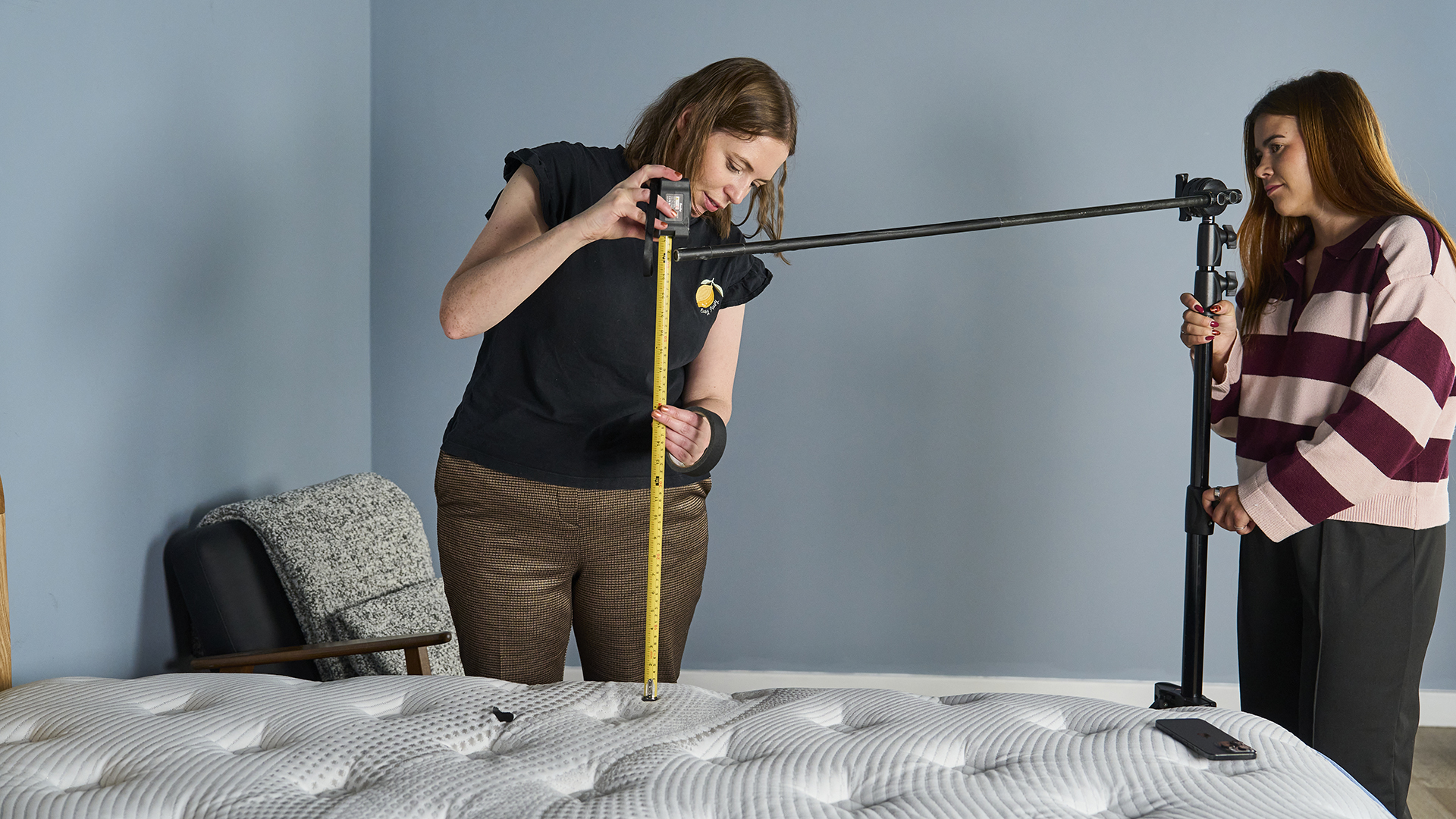 A close-up shot of two mattress testers performing a measurement on the Bear Elite mattress. One person, wearing a black shirt and brown trousers, is measuring the height from the mattress surface using a tape measure. The other person, wearing a striped sweater, is holding a tall, black pole that extends across the mattress for a drop test setup.