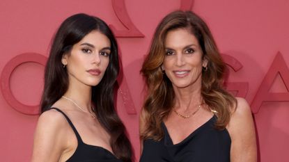 Kaia Gerber and Cindy Crawford stand together on a red carpet wearing coordinating black dresses and gold jewelry