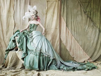 Woman in a green, ruffled ballgown, white feathers in her hair and a pink ribbon
