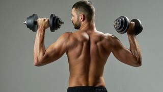 Man with back to camera topless holding two dumbbells over his shoulders and looking to the left against grey backdrop
