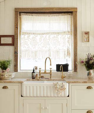 A white kitchen with fluted sink, brass taps, off-white shaker cabinets and shiplap walls, with wooden frame to large window