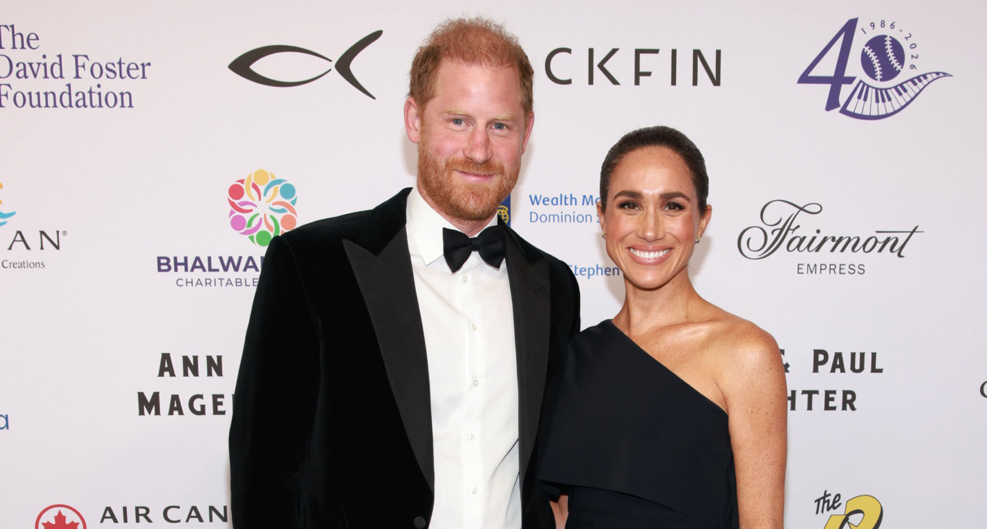 Prince Harry in a tux and Meghan Markle wearing a black gown on a red carpet