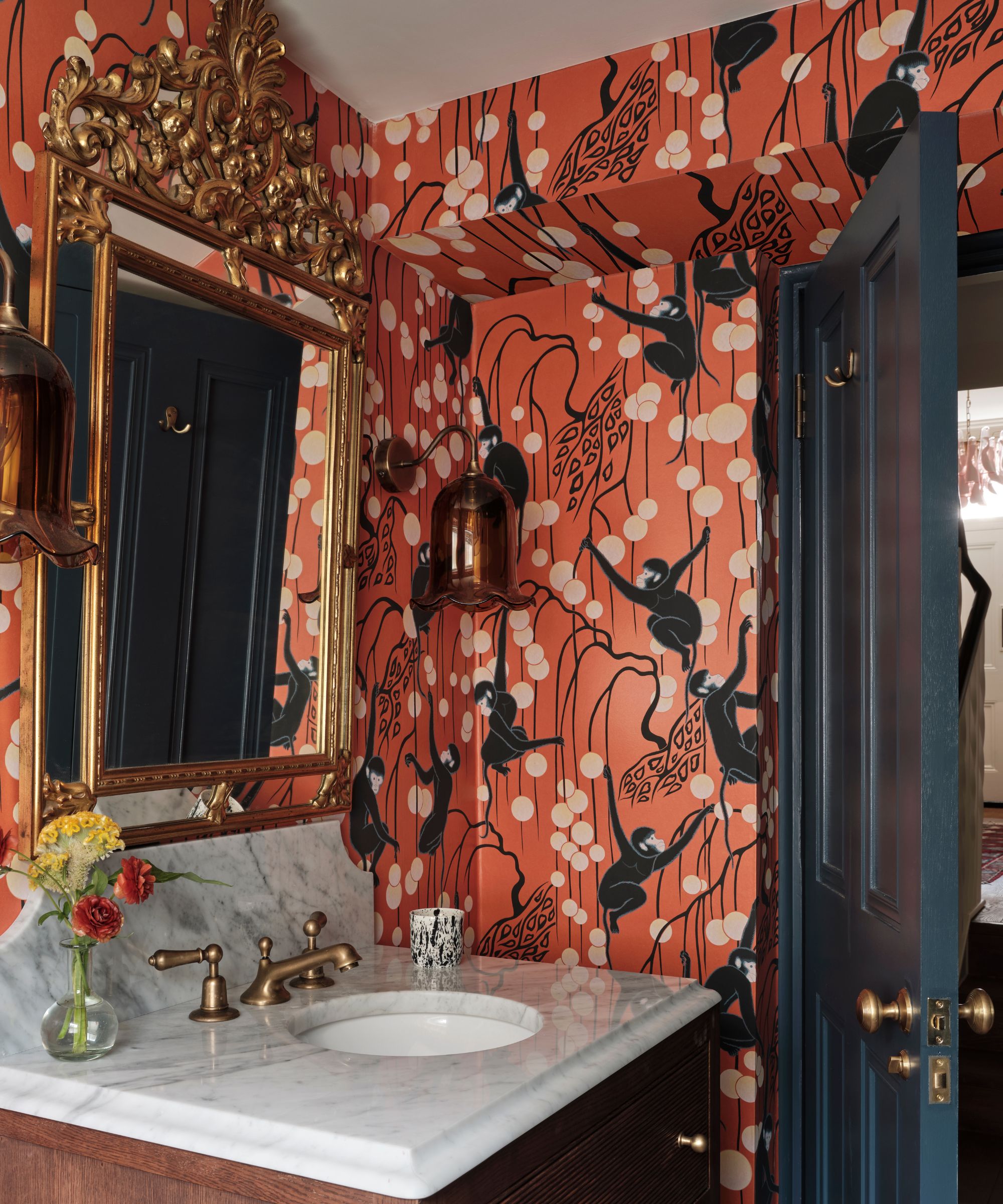 A small guest bathroom decorated with a red patterned wallpaper, a gold mirror, and a marble vanity