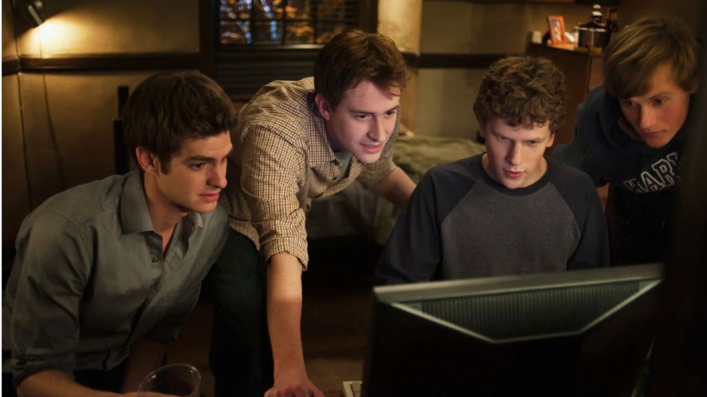 an image from the 2010 film The Social Network which shows four actors in a college dorm looking at a computer screen
