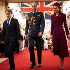 Prince George wears a suit, Prince William wears military uniform, Kate Middleton wears a dark red coat dress, as they walk in front of a Union Jack flag