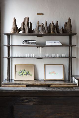 White rustic wall, dark wood shelving bar unit, two pictures in gold frames, drinking glasses on a shelf, books, Stone ornaments on the top shelf