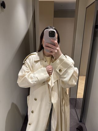 Woman in mirror wears cream trench coat and jeans.