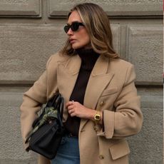 Woman wears camel wool jacket, black cashmere funnel neck jumper and blue jeans 