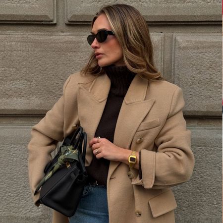 Woman wears camel wool jacket, black cashmere funnel neck jumper and blue jeans