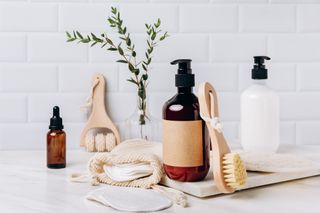 Bottles of shampoo, shower gel, moisturiser and serums, along with massage brushes and cotton pads in front of a white tiled wall.