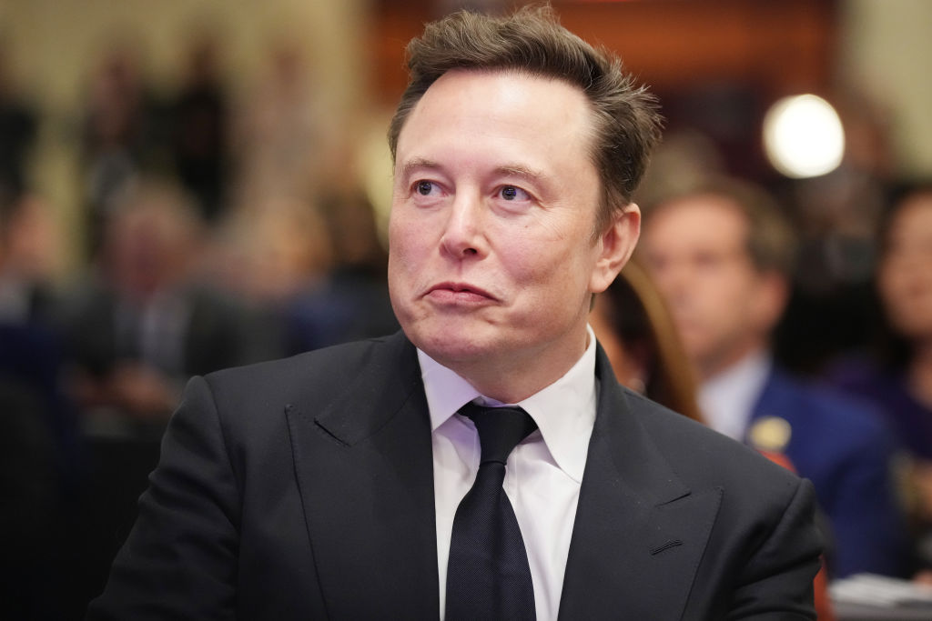 A man with dark hair wearing a black suit and tie looks to the left of the camera