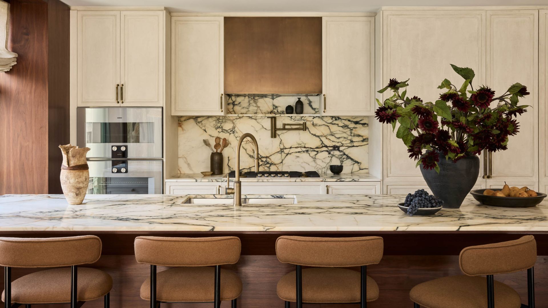 A bright kitchen with off-white cabinets, marble backsplash, a brass exhaust, in-built ovens, a marble island with brown kitchen stools, a plate of grapes, and a vase of flowers
