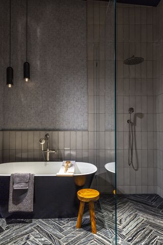 bathroom with zoned tub and shower areas/glass wall, grey walls, pale grey wall tiles, herringbone floor tiles, blue black, stool, towels, pendant