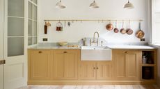 kitchen with white walls, yellow cabinetry, marble countertop, hanging rail with copper saucepans, trio of white pendant lights, white butler sink, brass faucet, wooden floorboards, doors on left, window on right