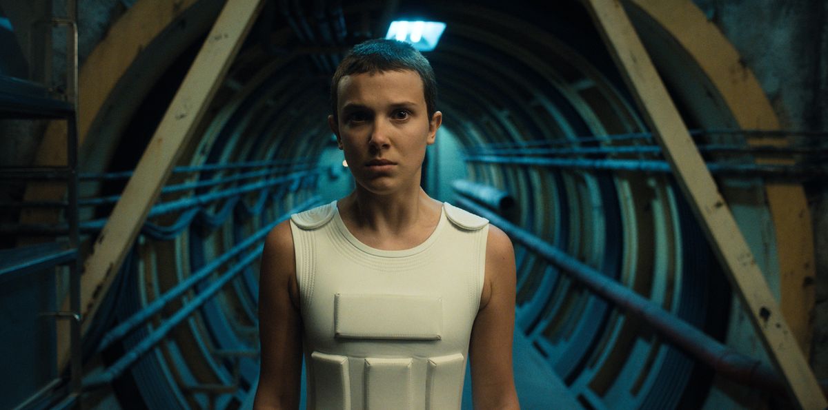 Eleven stands stoically in a corridor in an underground facility in Stranger Things 4