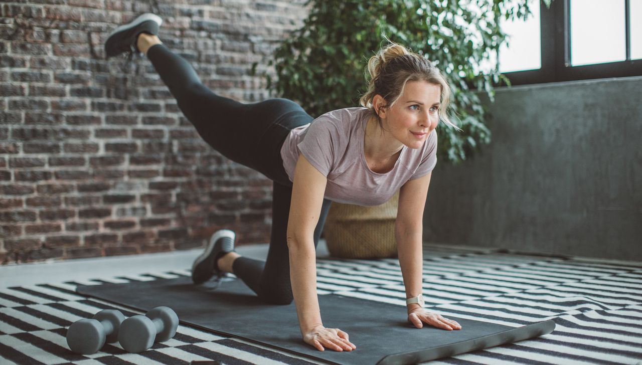 Woman at home exercising. She is on all fours with one leg raised and extended behind her. Her hair is in a bun and she is wearing black athletic leggings and a short-sleeve T-shirt.