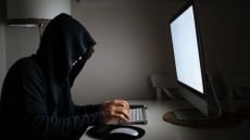 Hacker concept image showing young man with hood covering face typing on a keyboard in a dark room.
