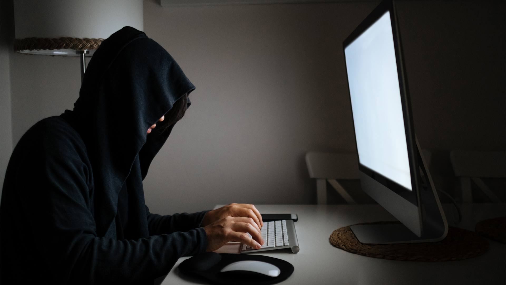 Hacker concept image showing young man with hood covering face typing on a keyboard in a dark room.