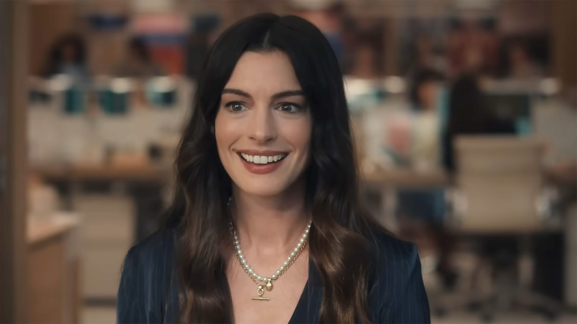 Anne Hathaway sits at a desk at Runway with a pearl necklace on in Devil Wears Prada 2.