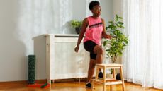 woman exercising in a pink vest and black shorts stepping up onto an indoor step in a living room setting. 