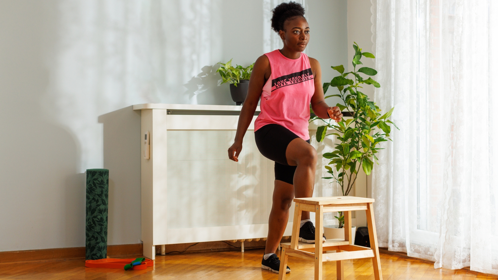 woman exercising in a pink vest and black shorts stepping up onto an indoor step in a living room setting.