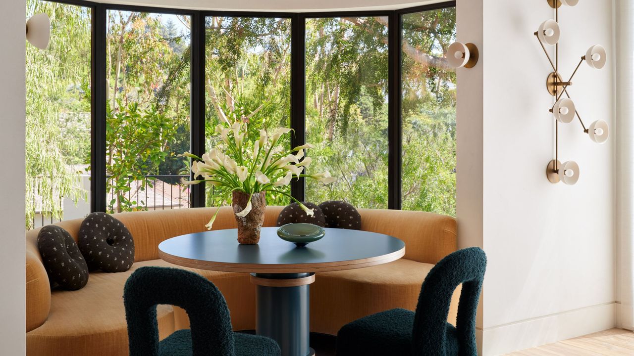 Image of a bright dining nook with a large, modern bay window that has a curved, orange bench seat, a round blue table, and two dark teal dining chairs. There are multiple sconce lights on the wall.
