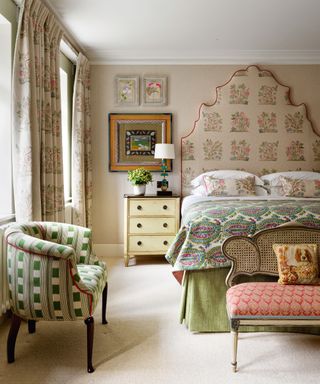 Bedroom with tall patterned headboard, patterned bedding, armchair and bench, patterned curtains on tall windows.