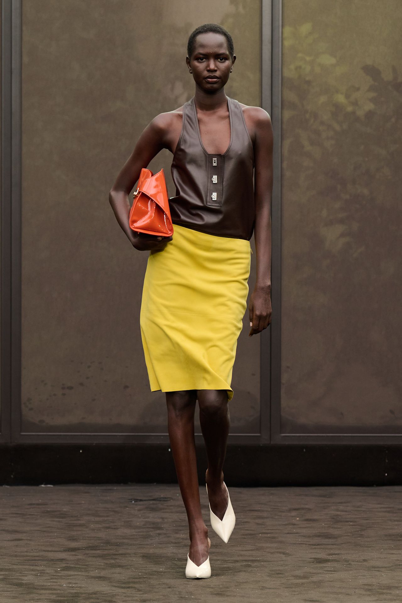 A Ferragamo model wearing a low scoop neck chocolate brown leather tank top with a citrus yellow suede midi skirt, white pointed heels, and a bright orange east-to-west clutch bag.