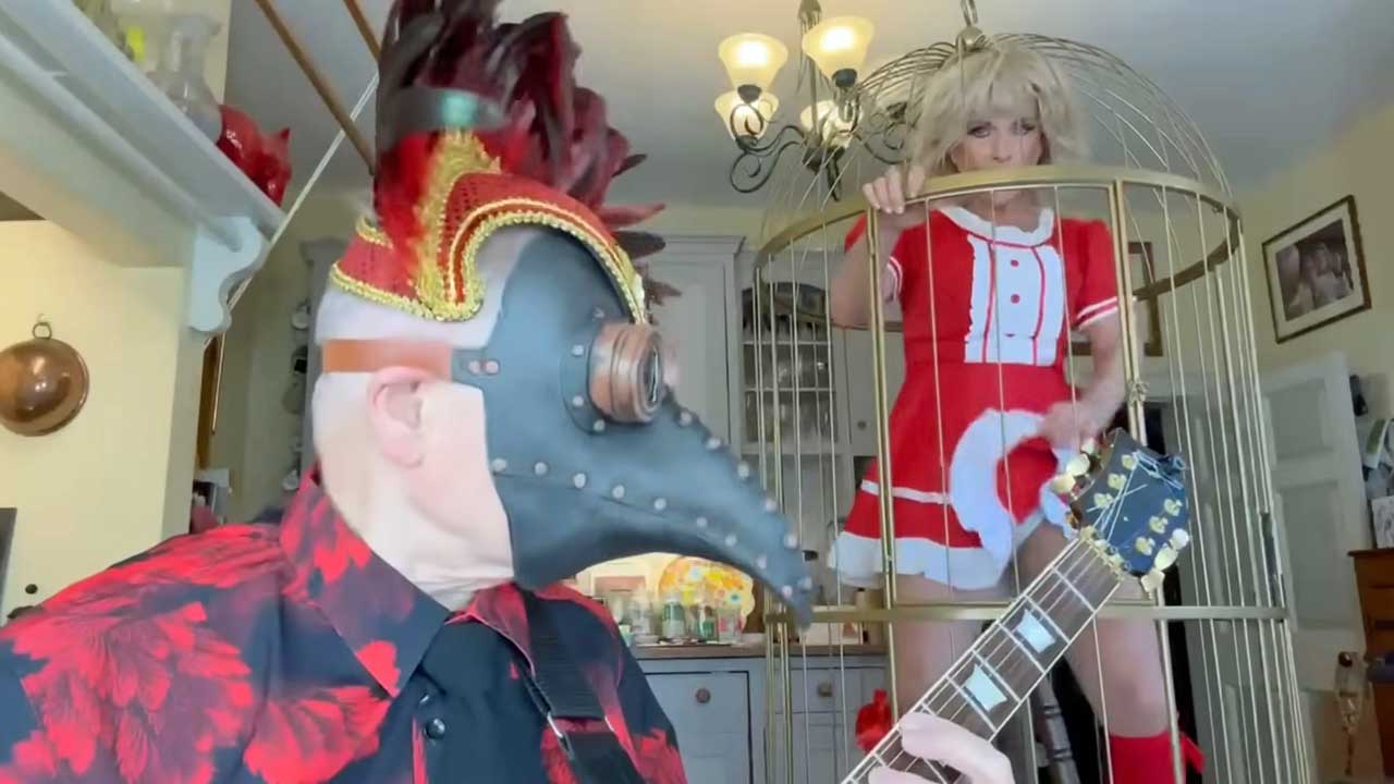 Robert Fripp wearing a plague doctor mask and a headress and Toyah dancing in a birdcage in their kitchen