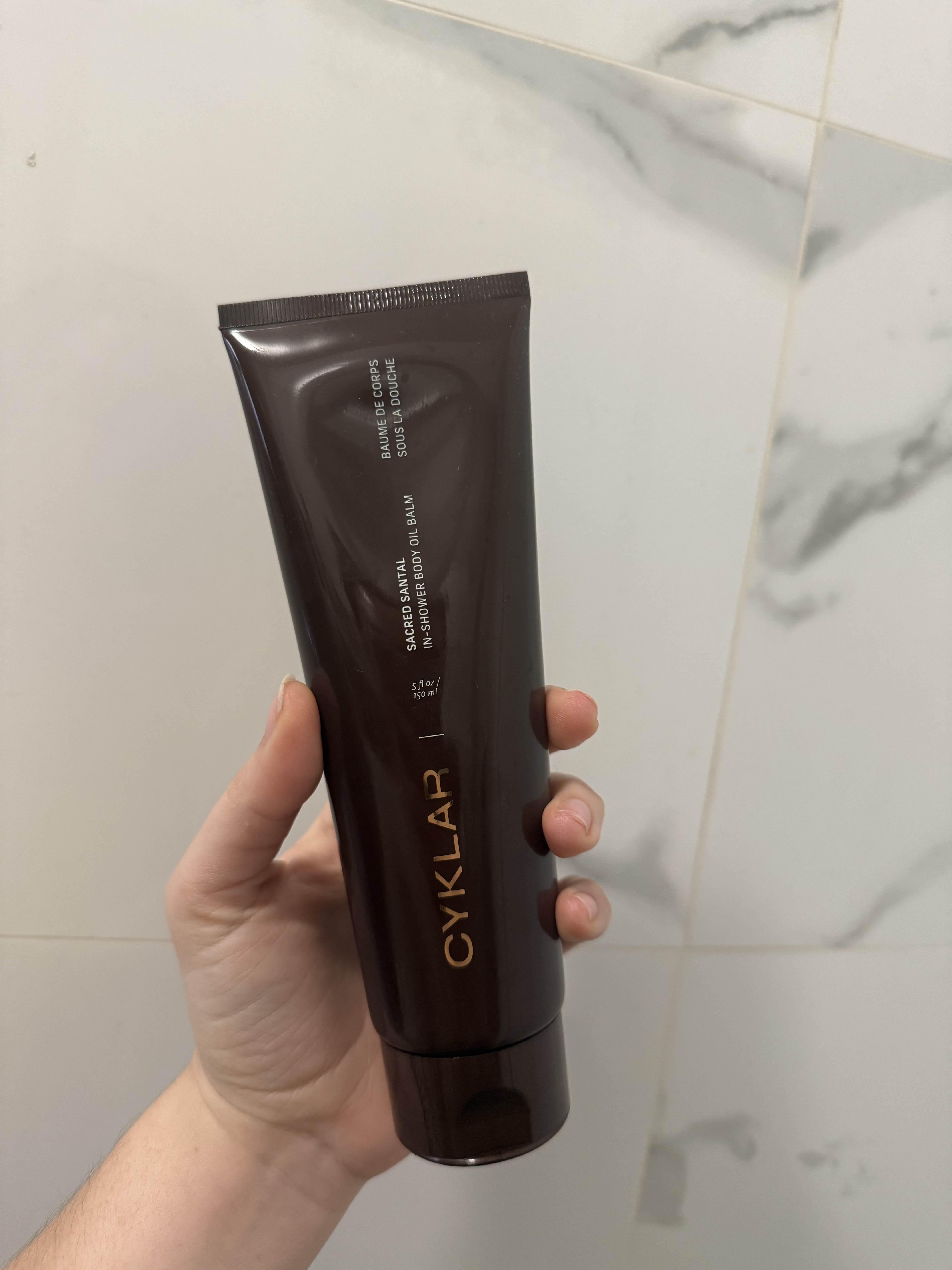 Hand holding up cyklar sacred santal in shower body oil balm against tile wall