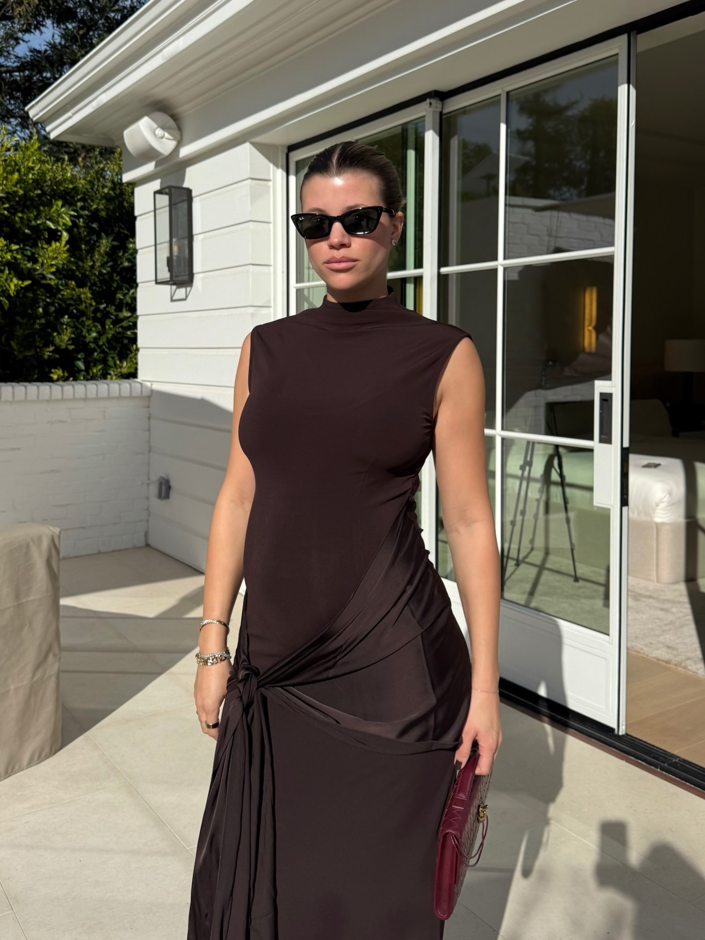 Sofia Richie Grainge wears a chocolate brown dress with a burgundy clutch bag and dark sunglasses.