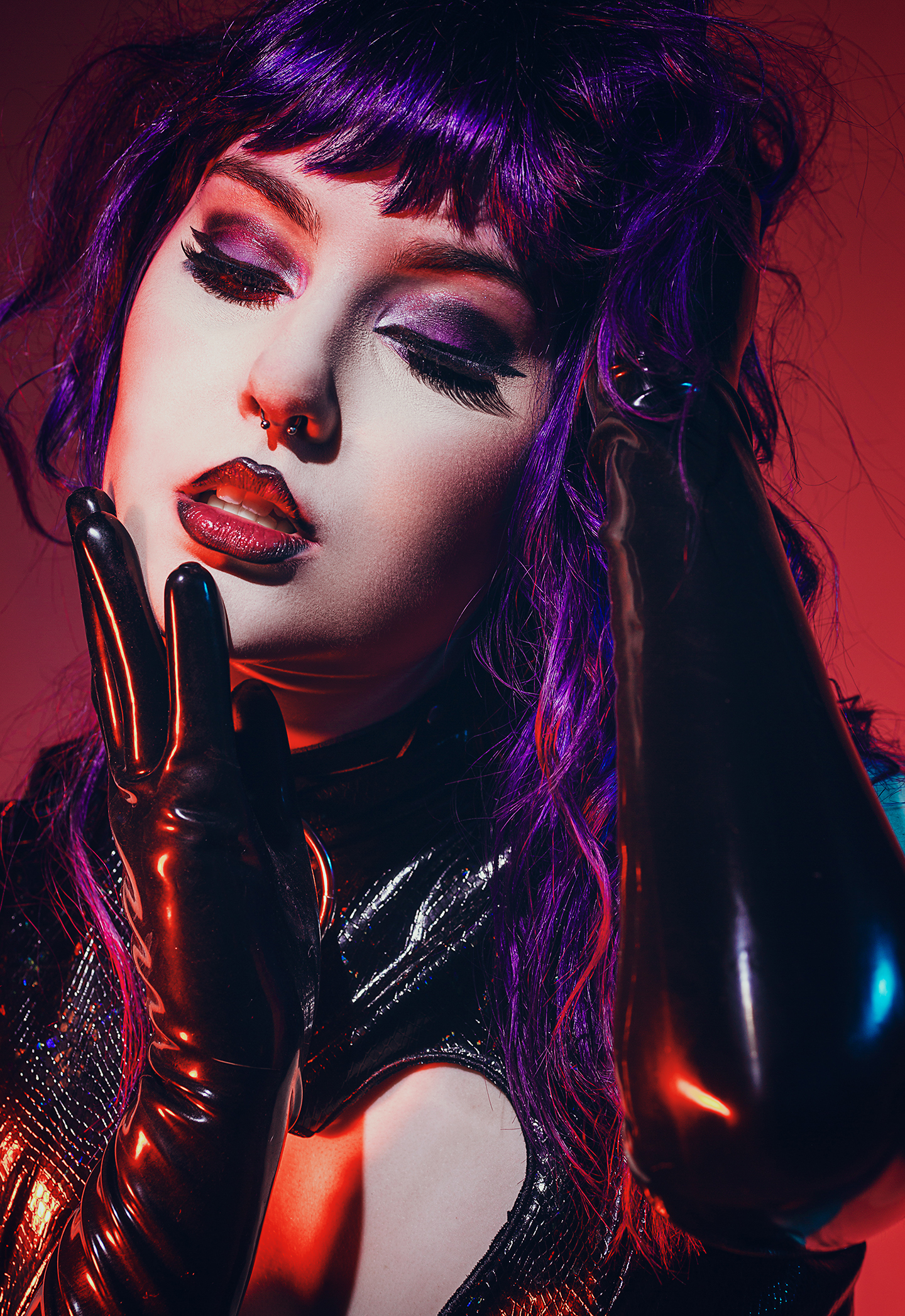 Woman with dark makeup and purple hair wearing a shiny black outfit and gloves, posed with eyes closed in dramatic lighting