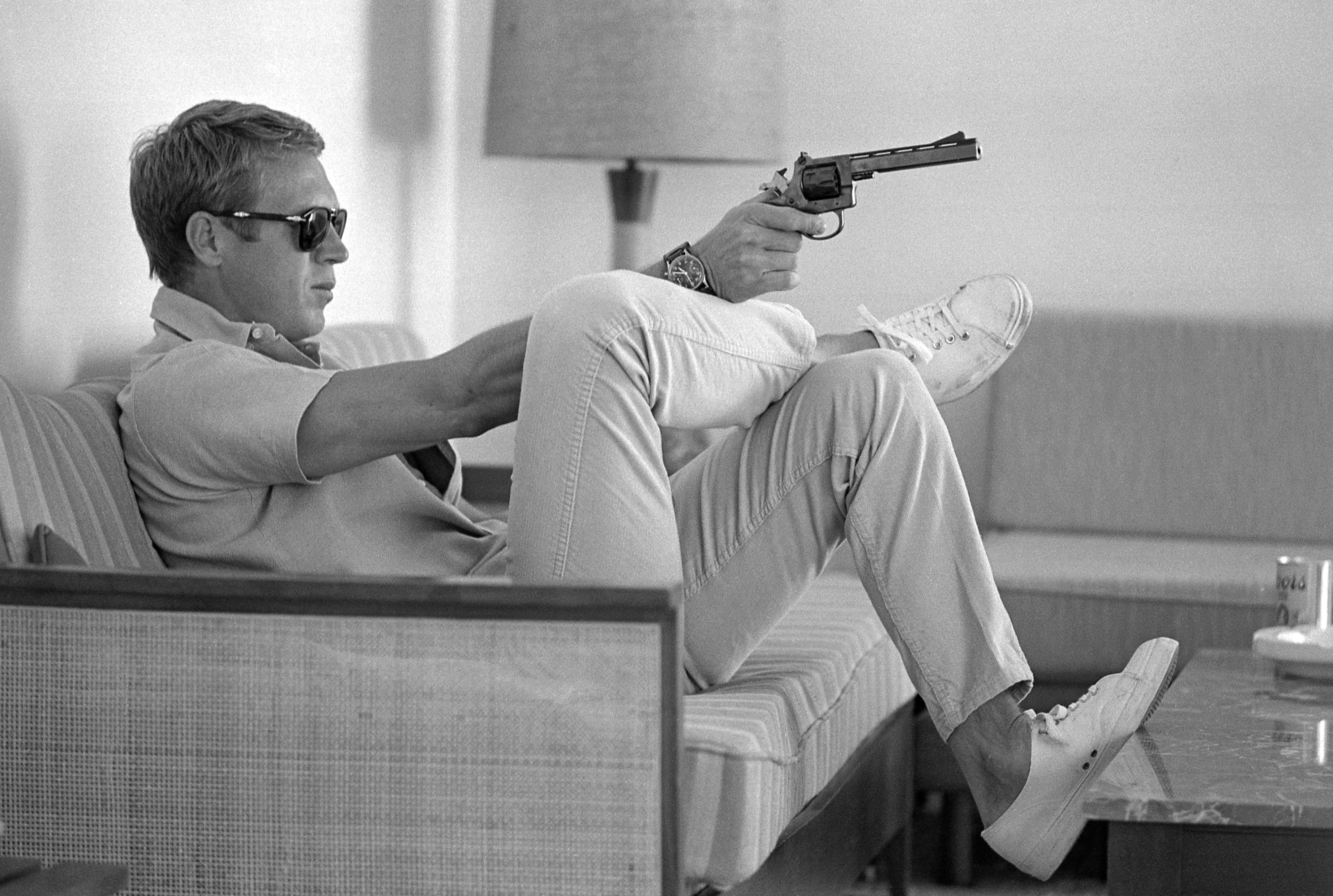 Steve McQueen photographed from the side. He's sitting on a sofa in white trousers and a polo shirt, pointing a gun at something or someone off camera