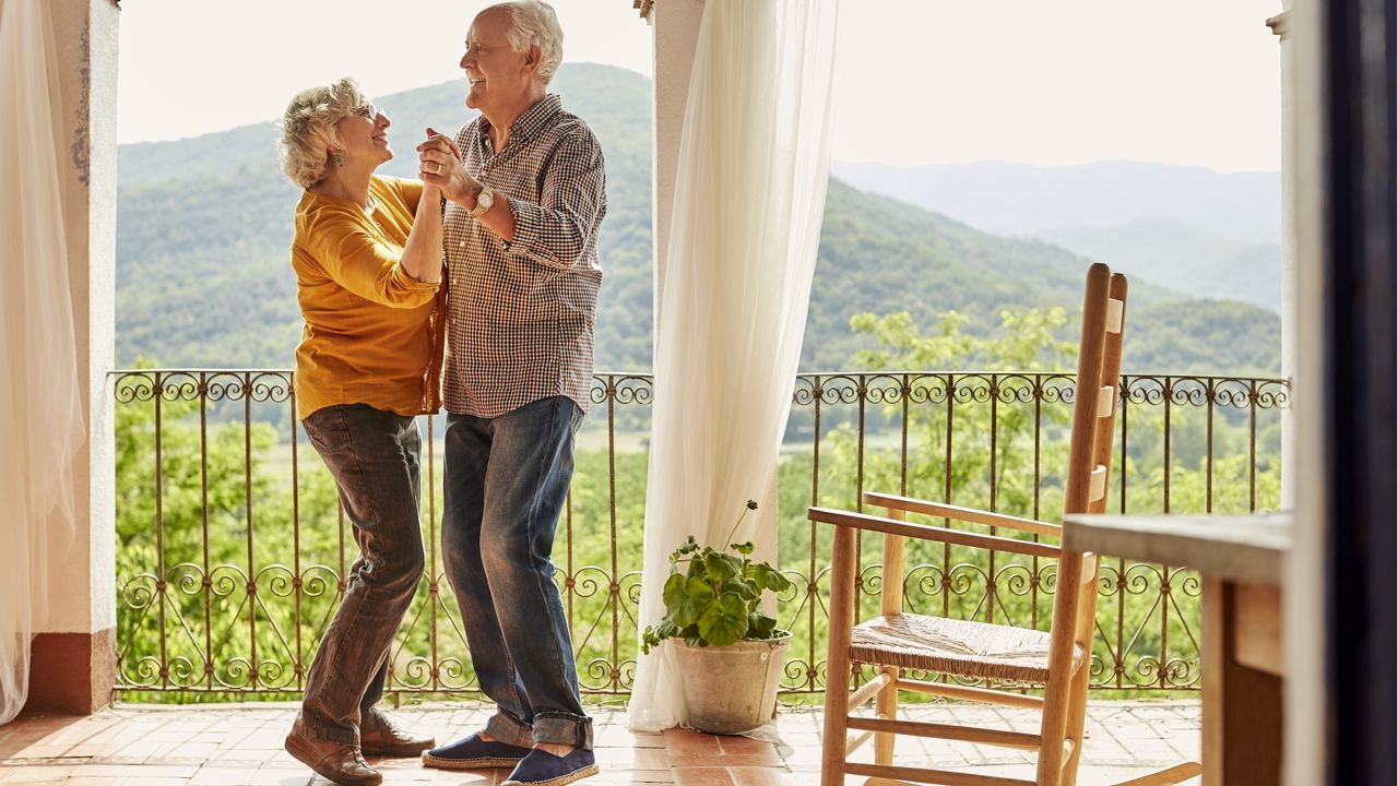 A happily retired couple dance together on their porch with a great mountain view behind them.