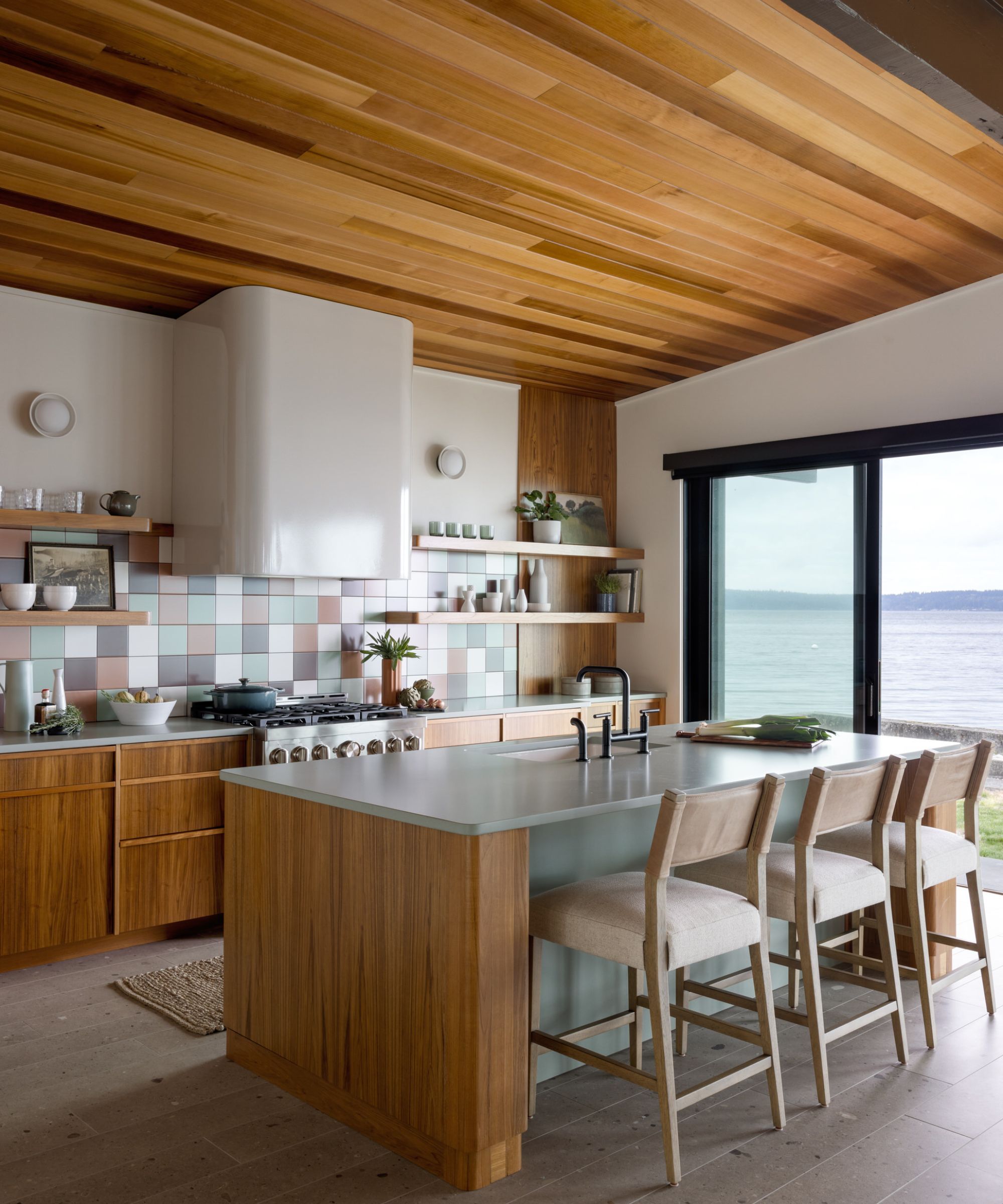 A modern beach house kitchen with a wood-clad ceiling, teak kitchen cabinets, off-white walls, square backsplash tiles in muted colors, a large island with light green satin countertops, and sliding doors with a sea view.