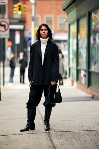 A woman standing on the sidewalk at New York Fashion Week wearing a black blazer, black pants, white turtleneck sweater, black riding boots, and a black Gucci bag.