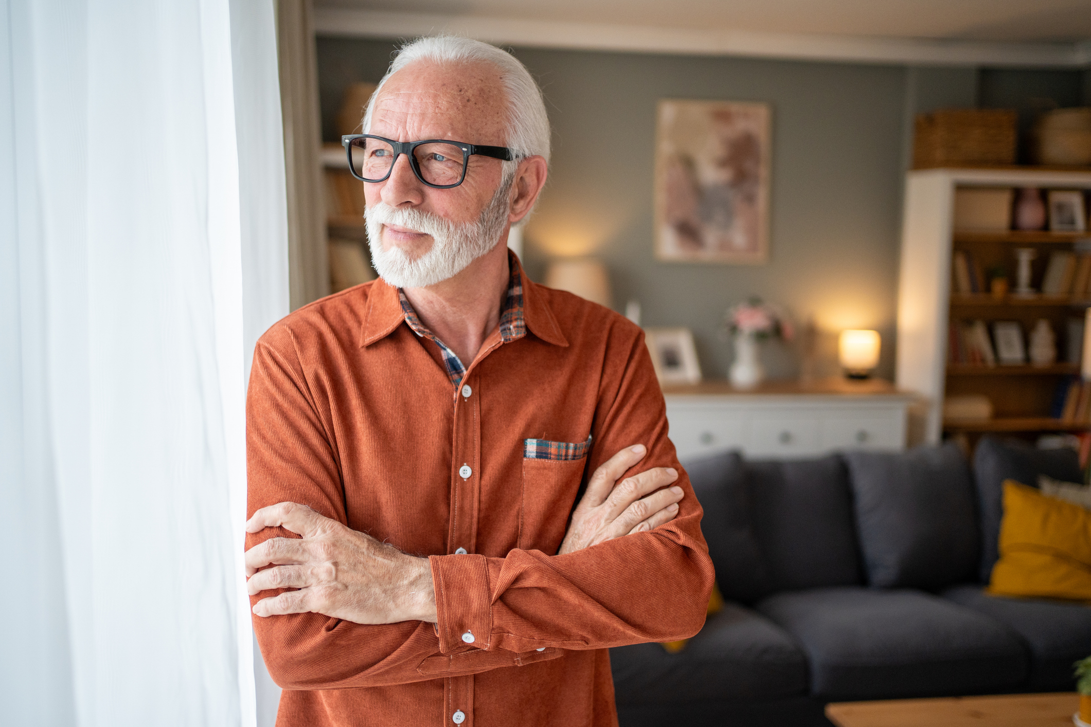 Thoughtful senior man standing by the window in a cozy living room, arms crossed and gazing pensively at the view, reflecting on life&amp;apos;s journey and cherished memories