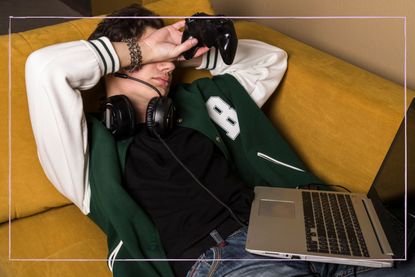 A boy or young man in headphones has fallen asleep on a couch with a controller in his hand and a laptop. The gamer had been playing all night and is now sleeping through the day