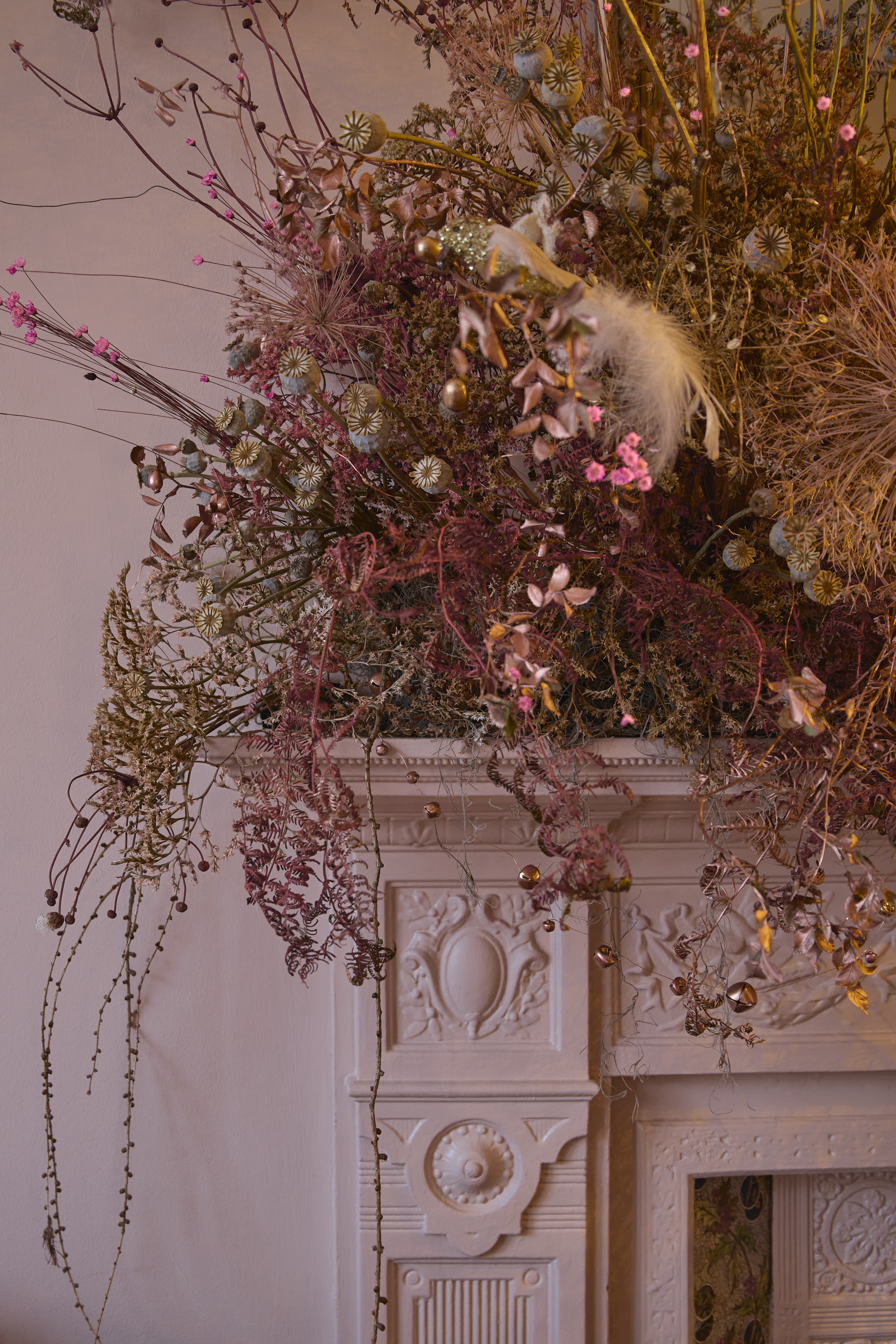 Left upper corner of a white, traditionally moulded fireplace. On top is large arrangement of dried florals in varying tones of muted pink, grey and brown.