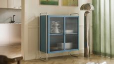 a light blue cabinet with reeded glass doors, a silver lamp, and light green curtains