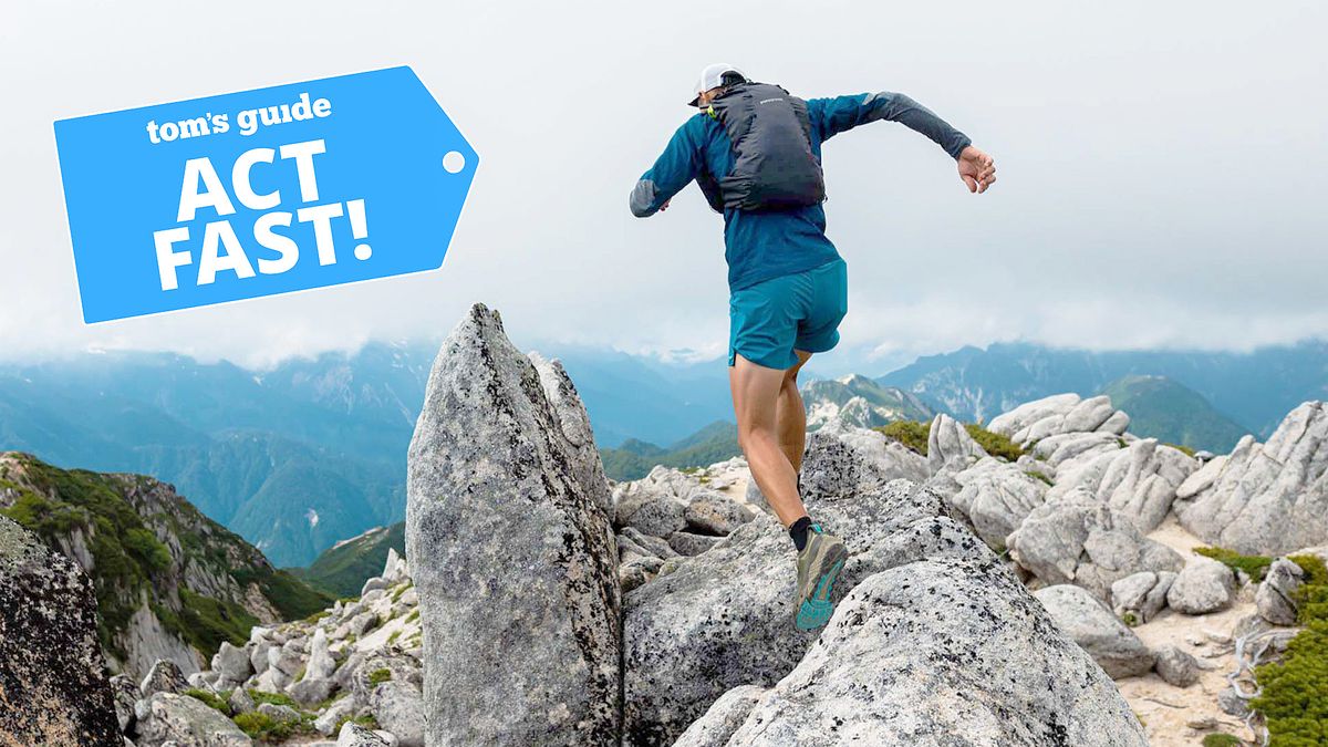 A trail runner dressed in blue cresting a mountain peak with other distance mountains in the background against a white cloudy sky.