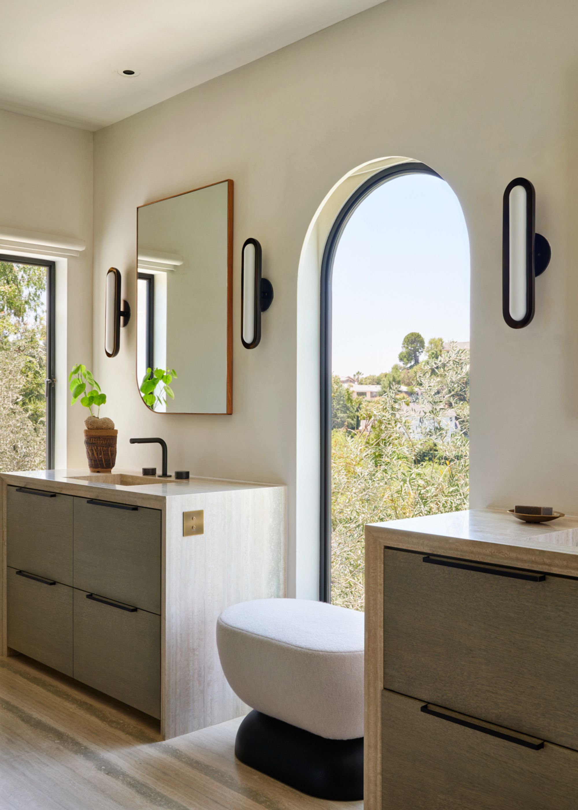 A his and hers bathroom with tubular sconces, hanging mirrors, and a two-toned bench by an arched window