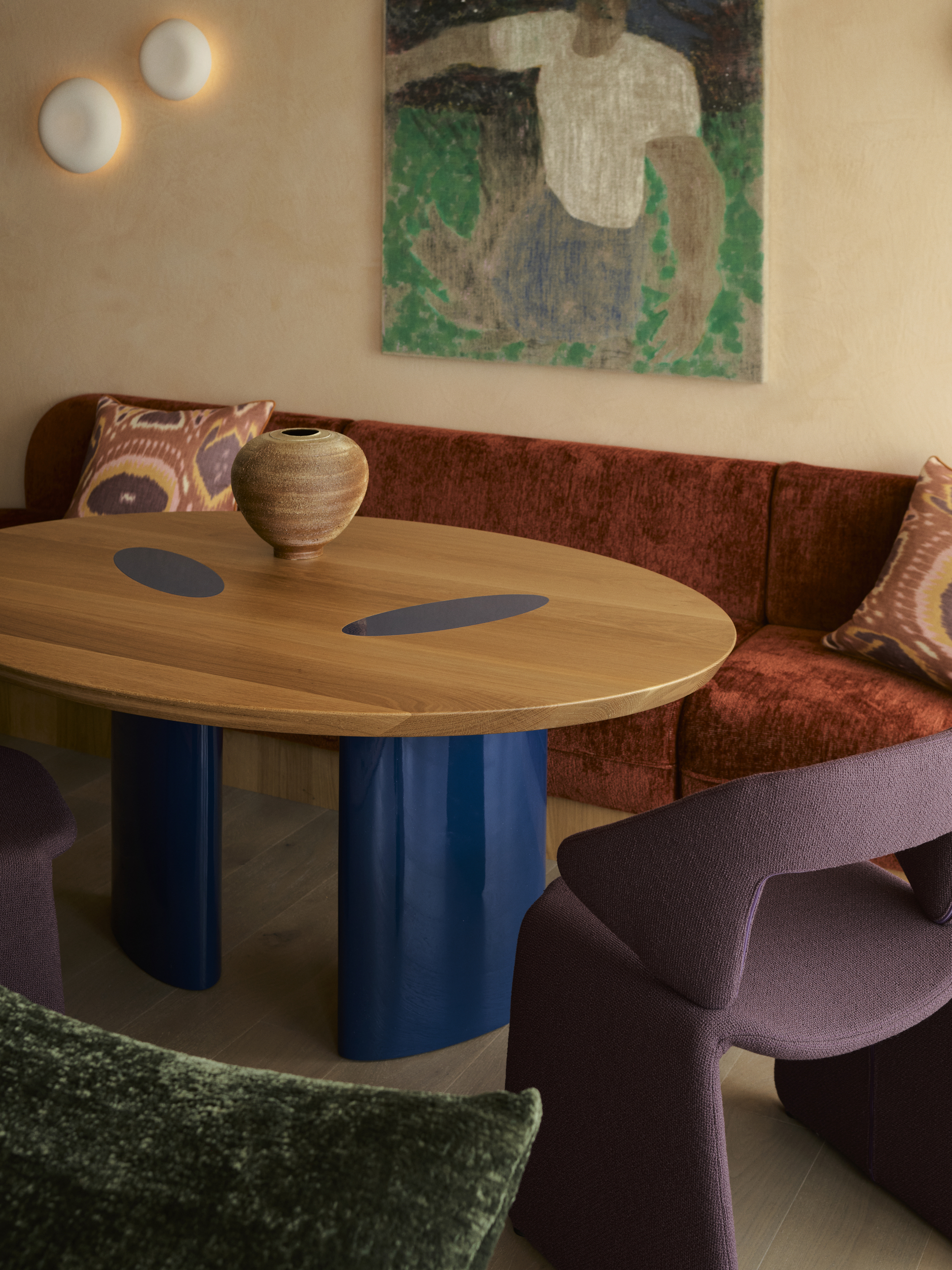 modern dining room with red banquette seat, timber table with blue legs, purple dining chairs, and timber floors, plaster walls, and large green artwork and wall sconce