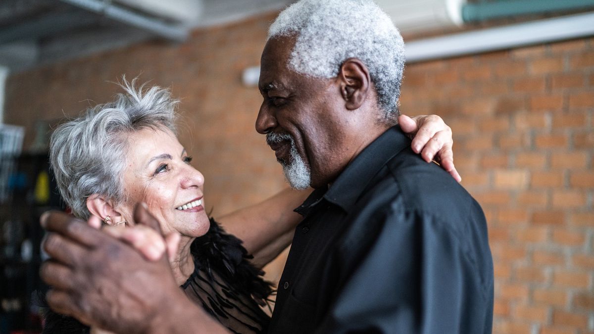 A senior couple, both with gray hair, are dancing a waltz in a dance hall.